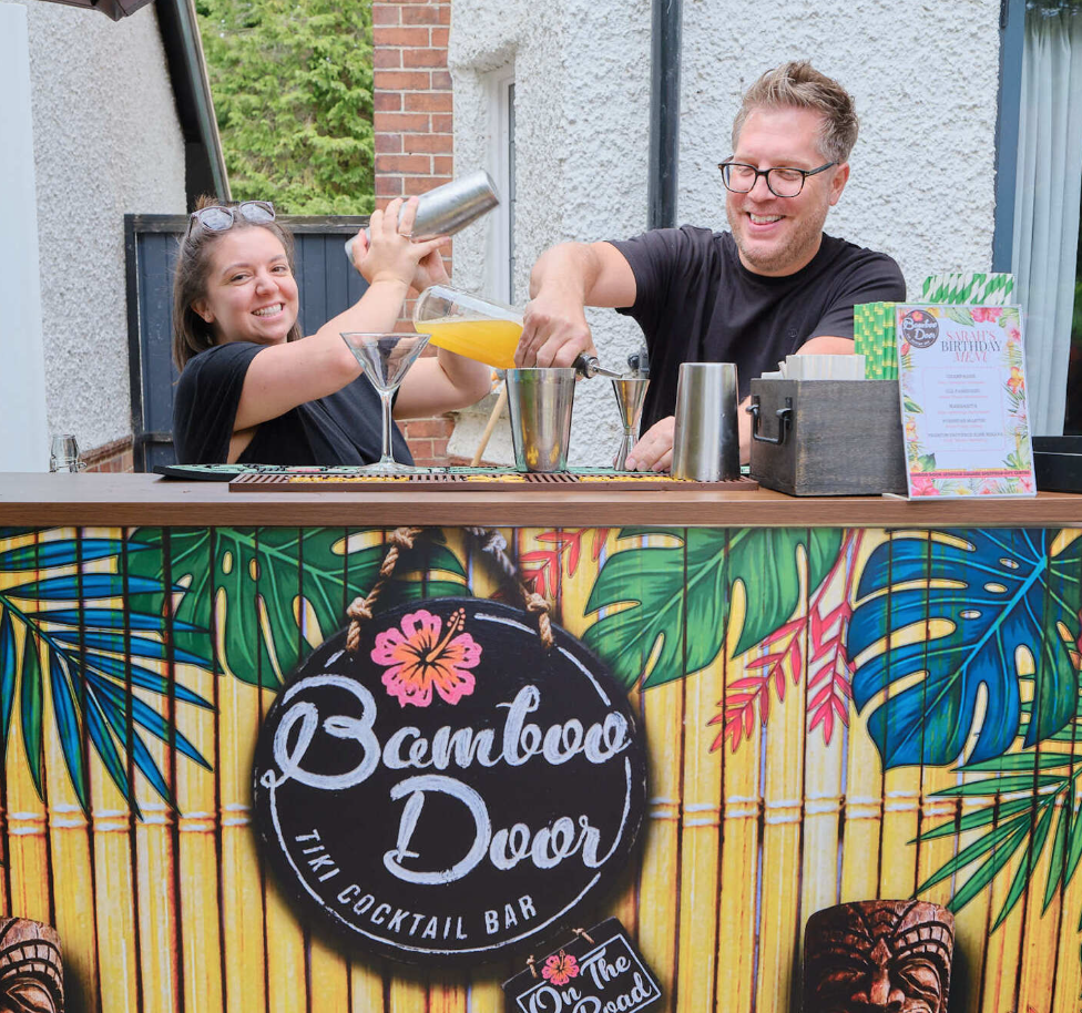 A professional photograph of two bamboo door on the road staff behind a mobile bar that is hired for a party, making two tiki bar cocktails